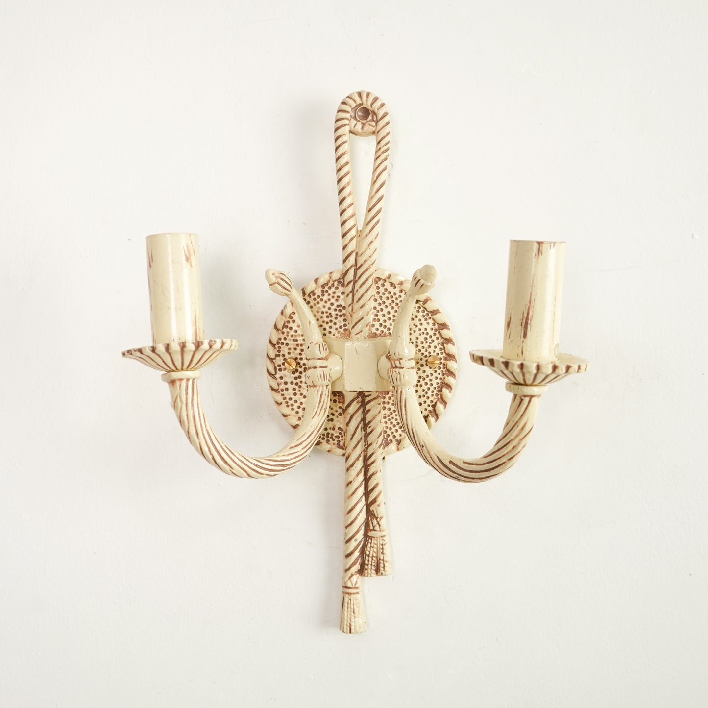 A two-light wall sconce with a distressed cream and brown finish, featuring twisted rope-style arms and a circular textured backplate, shown against a plain white wall.