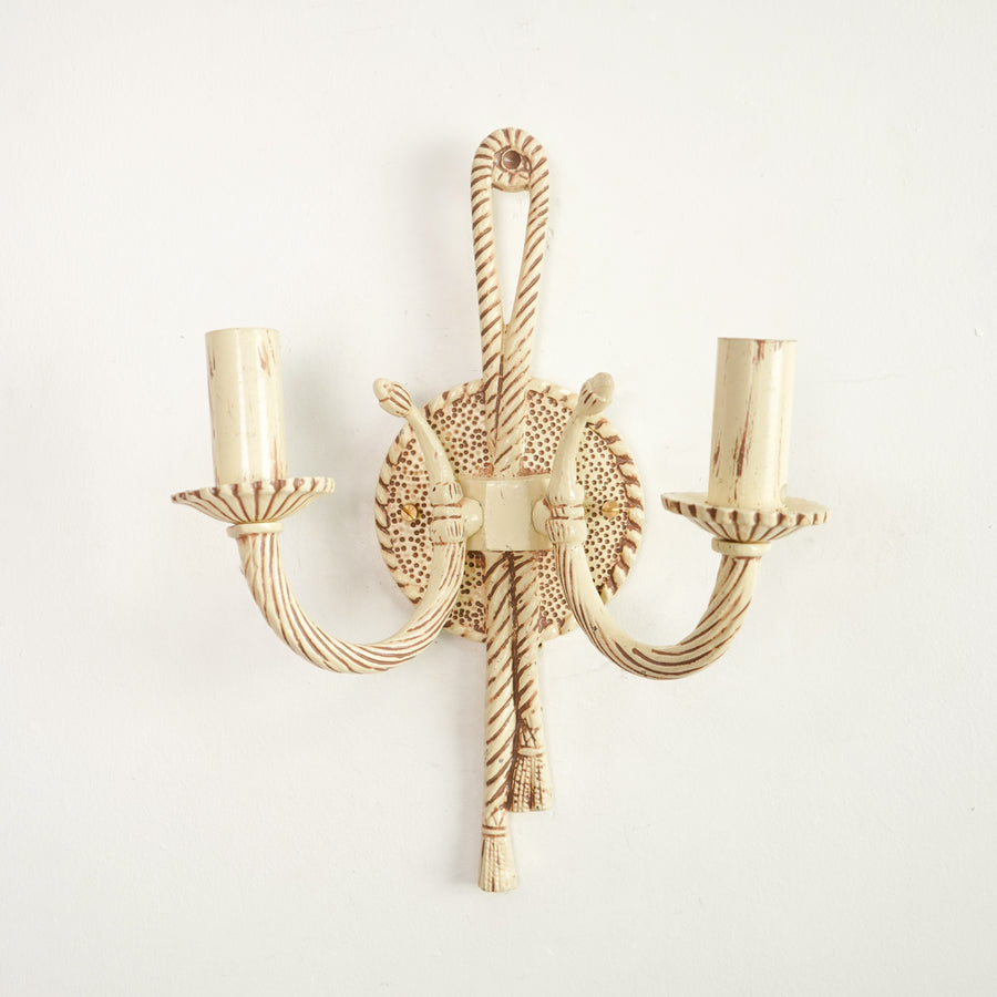 A two-light wall sconce with a distressed cream and brown finish, featuring twisted rope-style arms and a circular textured backplate, shown against a plain white wall.