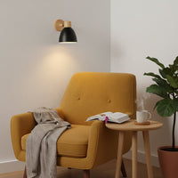 Modern wall light with black metal shade and wooden mount placed above lounge chair creating warm reading corner lighting for living room decor.