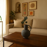 Modern living room setting with a textured grey jug vase placed on a wooden table in front of a beige sofa and framed artwork creating a warm decorative atmosphere.