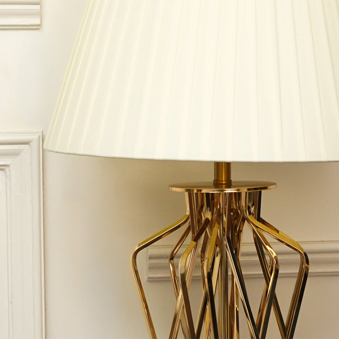 A close-up of a table lamp with a pleated off-white fabric shade and a polished gold geometric metal base, set against a cream-colored wall with decorative molding.