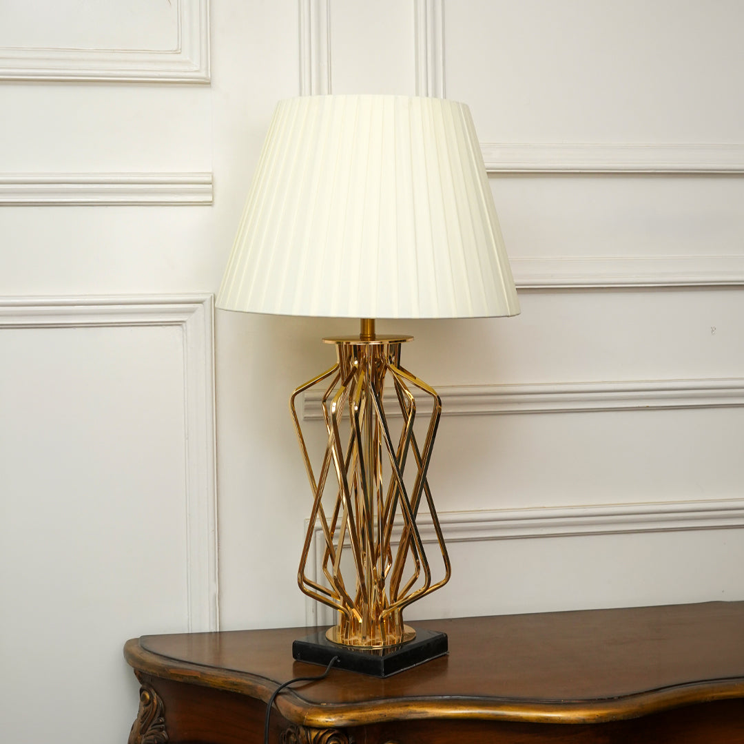 A modern table lamp with a pleated off-white shade and a gold geometric wireframe base on a black square pedestal, set on a dark wood console table against a white paneled wall.
