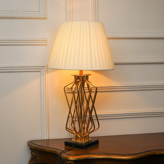 A table lamp with a pleated off-white shade and a gold geometric wireframe base on a dark square pedestal, set on a wooden console table against a cream paneled wall.
