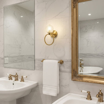 Gold metal wall light with textured glass globe illuminating marble tiled bathroom vanity with mirror towel ring and classic sink fixtures.