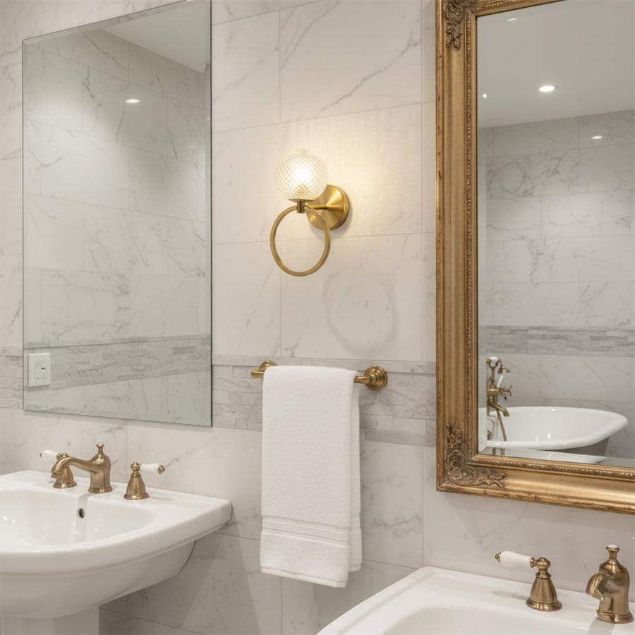 Gold metal wall light with textured glass globe illuminating marble tiled bathroom vanity with mirror towel ring and classic sink fixtures.