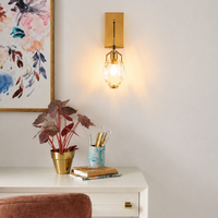 A modern wall-mounted sconce with a gold rectangular backplate and a clear faceted glass shade glowing with warm light, installed on a white wall above a white desk with a plant and books.