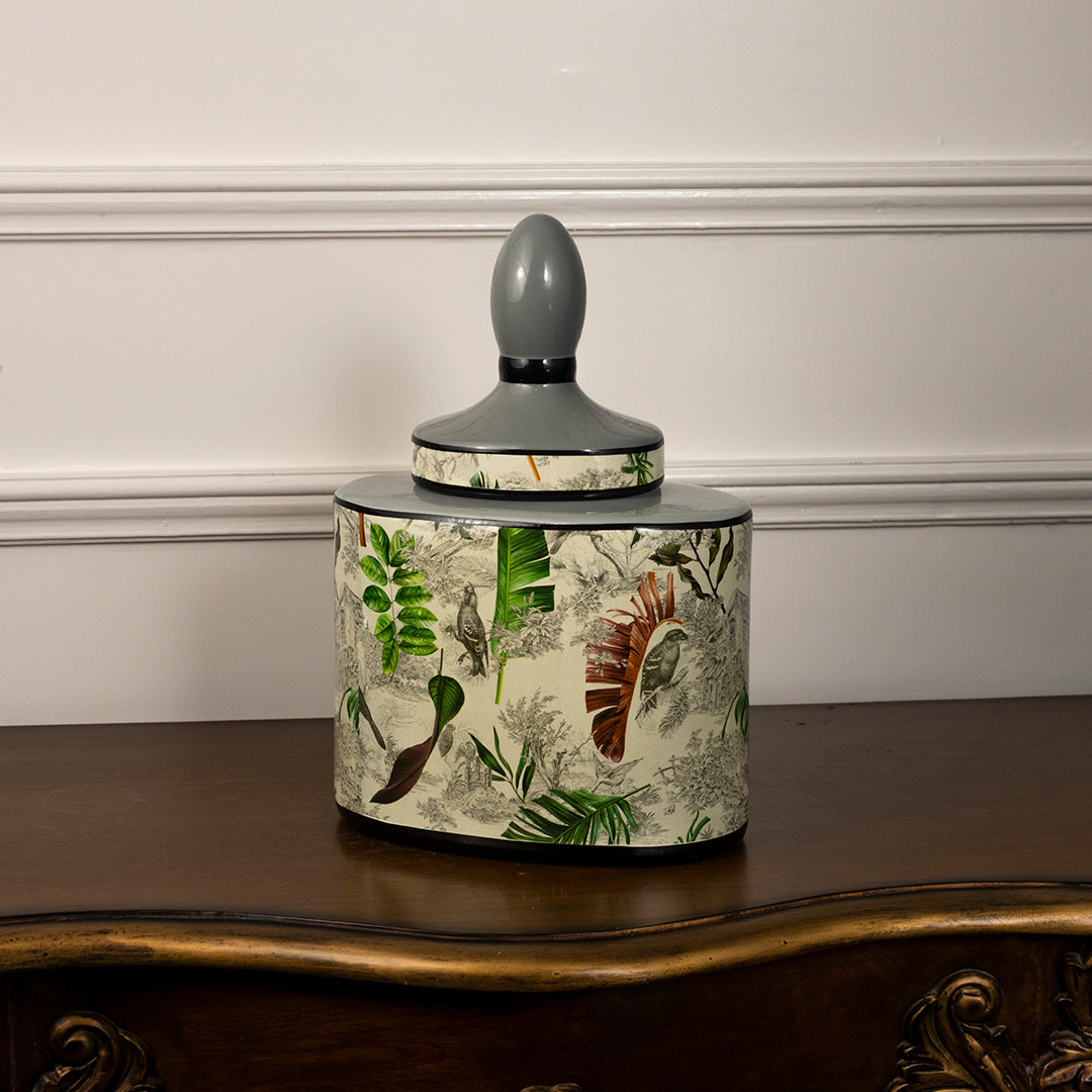 Ceramic storage jar with botanical leaf artwork and grey lid placed on wooden table against neutral wall background enhancing elegant nature inspired home decor appeal.