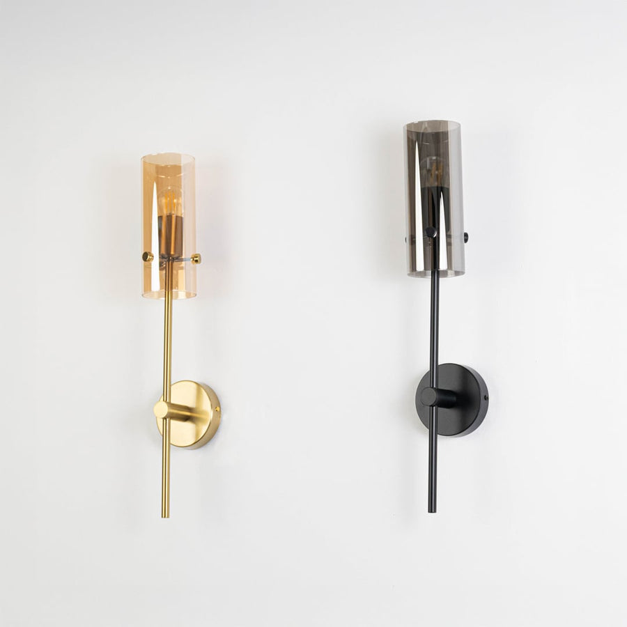 Pair of modern wall sconces in black and gold finish with cylindrical glass shades displayed against plain white background view.