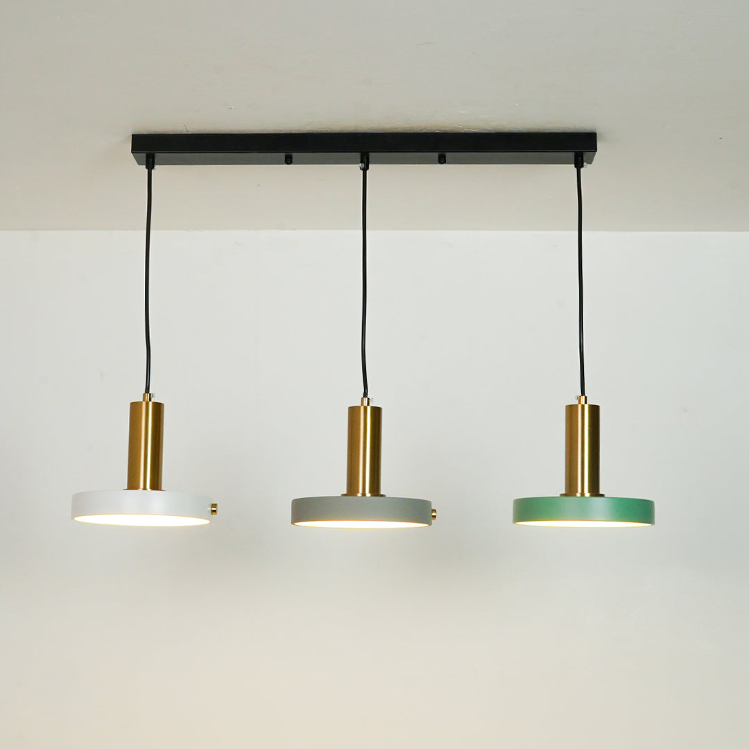 Three-light linear ceiling pendant fixture with a black horizontal bar, black cords, and three round shades in white, gray, and green with brushed gold centers, shown against a plain off-white wall.