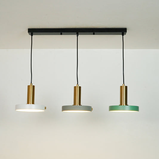 Three-light linear ceiling pendant fixture with a black horizontal bar, black cords, and three round shades in white, gray, and green with brushed gold centers, shown against a plain off-white wall.