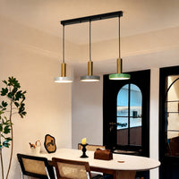 A modern three-light linear pendant chandelier hanging above a dining table in a real interior room. The fixture has a black rectangular ceiling mount, three black hanging cords, cylindrical gold-toned shades, and three round white/gray diffusers, with one light showing a green glow.