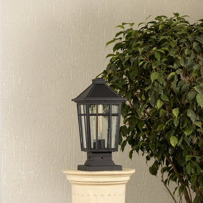 Black outdoor pillar light with ribbed glass Light On placed next to a lush green plant Lifestyle image for modern patio decor.