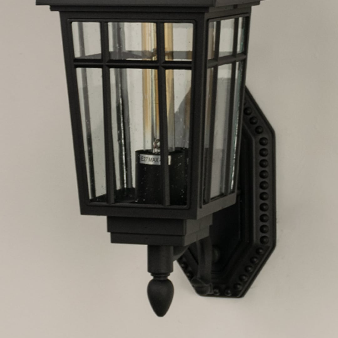 Closeup of a black outdoor lantern showing warm light shining through clear glass panels revealing textured reflections and classic lantern craftsmanship.