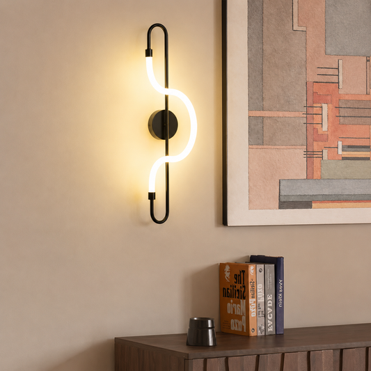 A modern wall-mounted light fixture with a black curved metal frame and a glowing white S-shaped curved tube, mounted on a beige wall above a wooden console table with books and a framed abstract print.