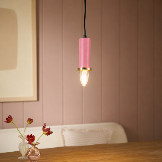 A pink cylindrical pendant light with a gold accent ring and a glowing bulb, hanging above a wooden table in a softly lit interior with a pink paneled wall and small vases in the foreground.