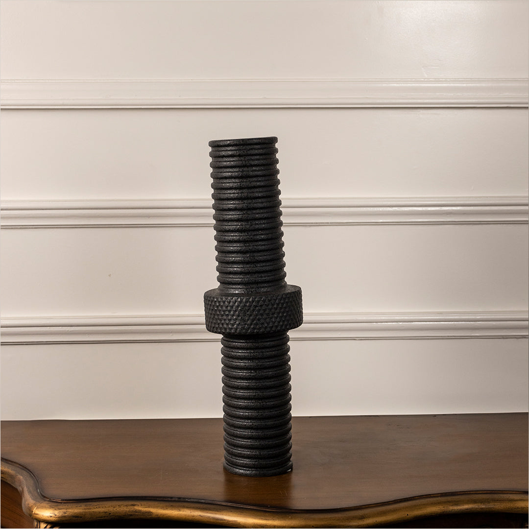 Black ribbed ceramic vase with stacked cylindrical design placed on wooden table against neutral wall background showcasing bold modern industrial decor styling.
