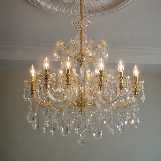 A multi-arm crystal chandelier with gold-toned metal arms and hanging clear teardrop prisms, shown illuminated against a neutral interior wall and ceiling.