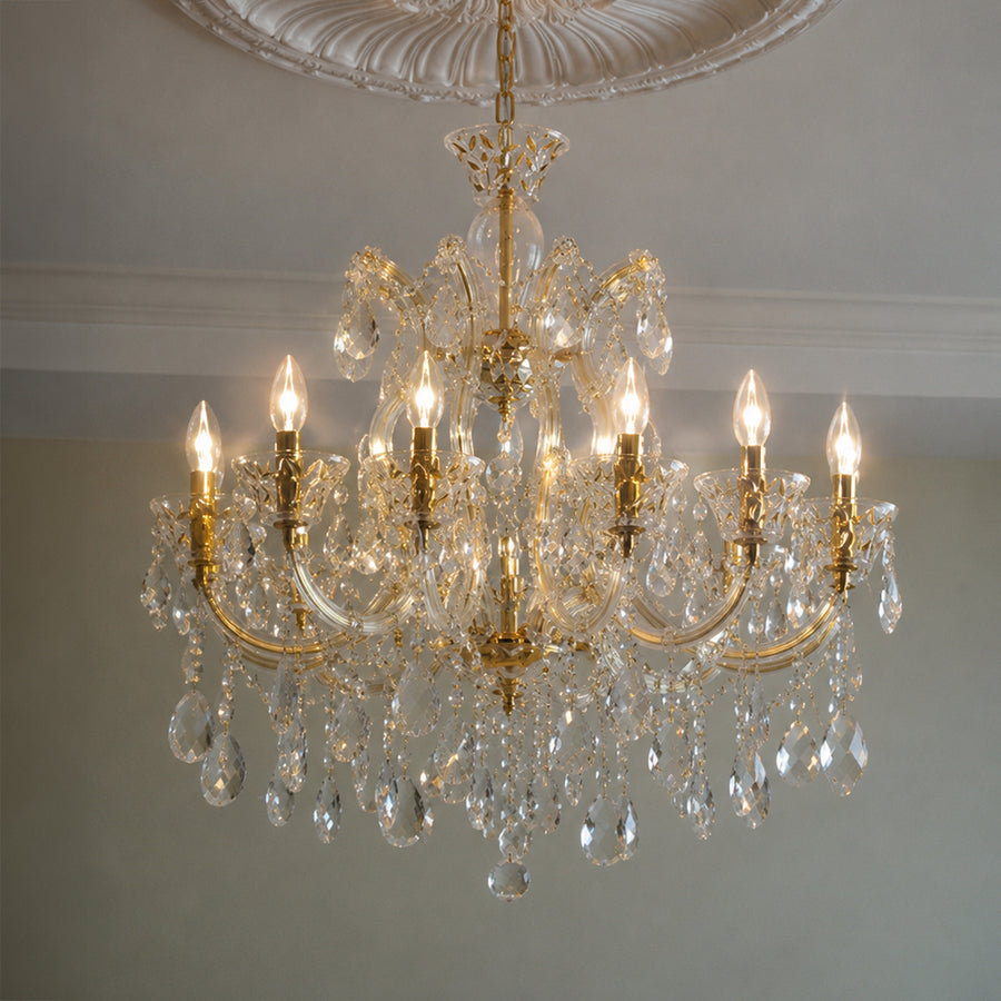 A multi-arm crystal chandelier with gold-toned metal arms and hanging clear teardrop prisms, shown illuminated against a neutral interior wall and ceiling.