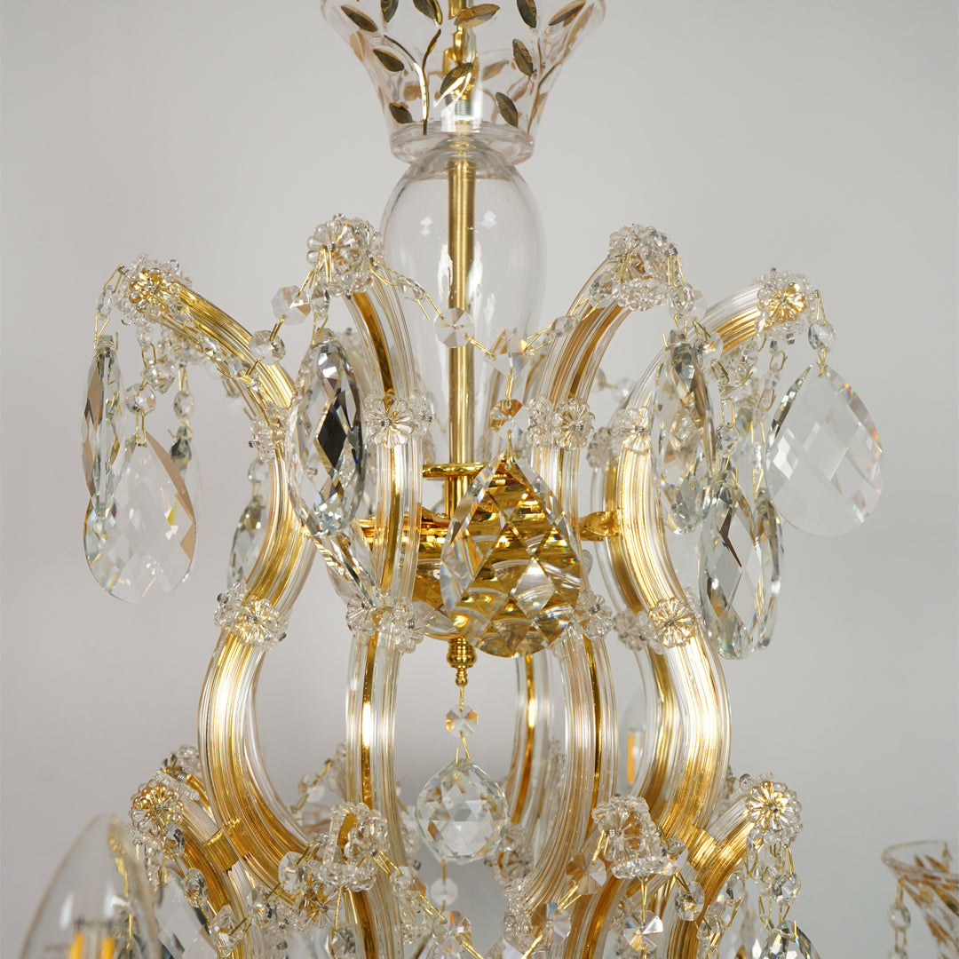 Gold and clear crystal chandelier detail with curved gold metal arms, faceted crystal pendants, and a glass globe in the center against a light gray background.