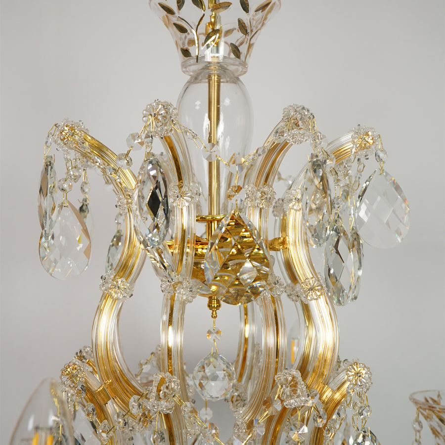 Gold and clear crystal chandelier detail with curved gold metal arms, faceted crystal pendants, and a glass globe in the center against a light gray background.