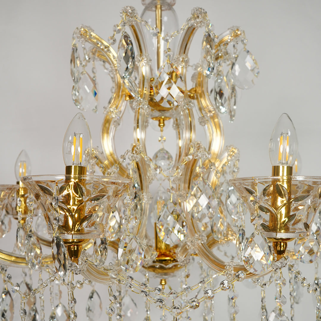 An ornate six-arm chandelier featuring a polished gold-finished metal frame adorned with clear crystal prisms and hanging teardrop drops, shown against a neutral light gray background.