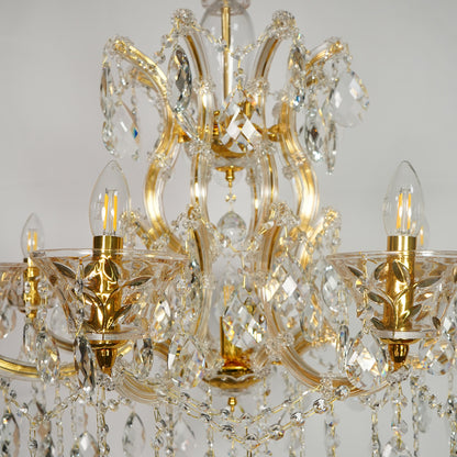 An ornate six-arm chandelier featuring a polished gold-finished metal frame adorned with clear crystal prisms and hanging teardrop drops, shown against a neutral light gray background.