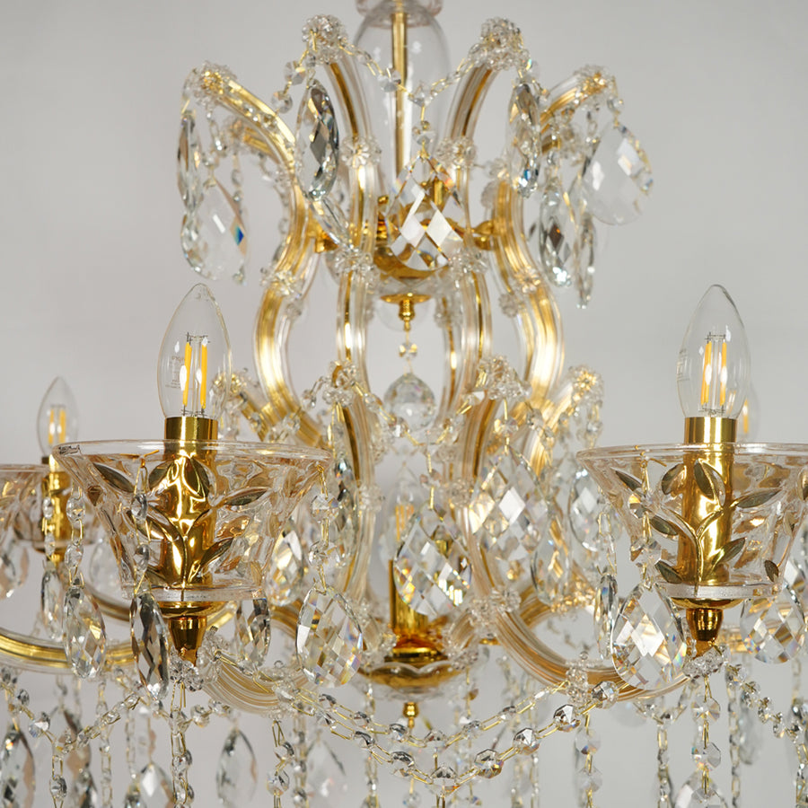 An ornate six-arm chandelier featuring a polished gold-finished metal frame adorned with clear crystal prisms and hanging teardrop drops, shown against a neutral light gray background.