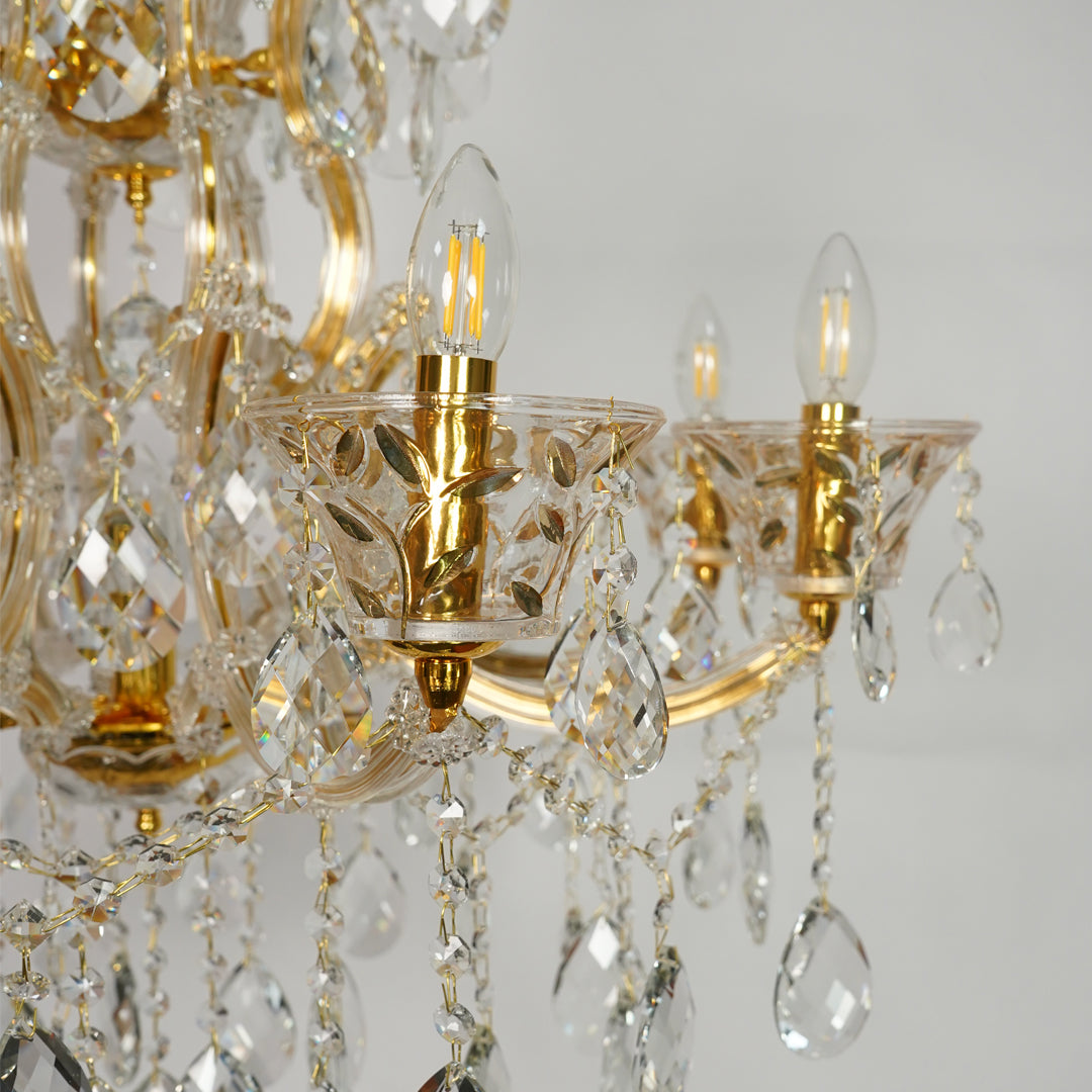 Gold and clear crystal chandelier close-up showing ornate gold metal arms, faceted crystal glass cups, hanging teardrop pendants, and lit candle-style bulbs against a light gray background.