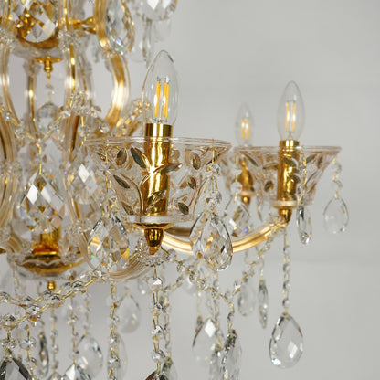 Gold and clear crystal chandelier close-up showing ornate gold metal arms, faceted crystal glass cups, hanging teardrop pendants, and lit candle-style bulbs against a light gray background.