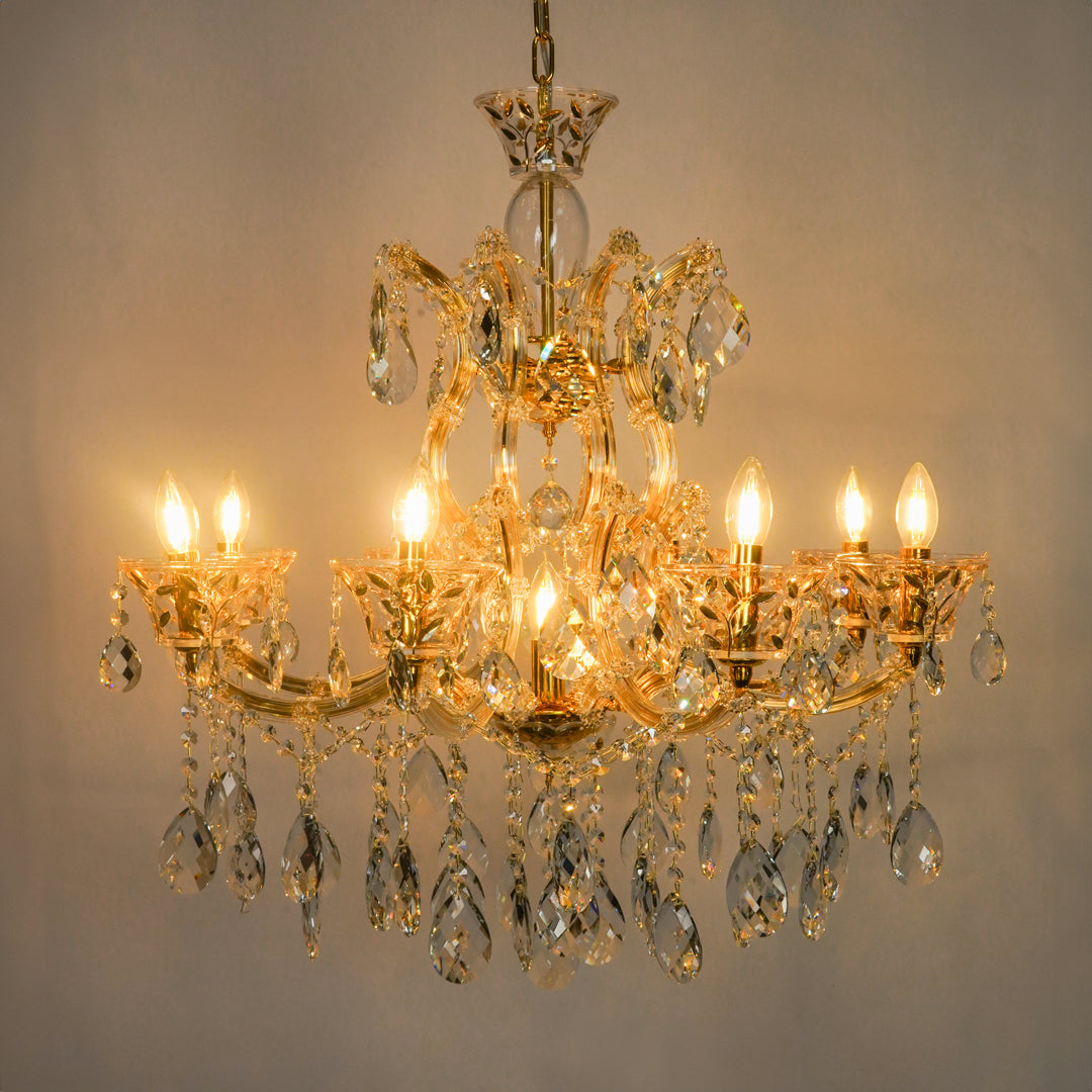 A multi-tier gold chandelier with clear crystal droplets and lit candle-style bulbs, photographed against a plain light gray wall.