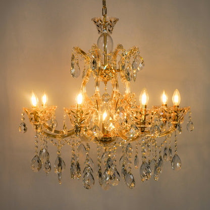 A multi-tier gold chandelier with clear crystal droplets and lit candle-style bulbs, photographed against a plain light gray wall.