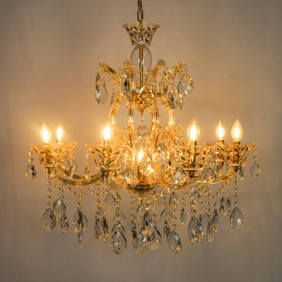 A multi-tier gold chandelier with clear crystal droplets and lit candle-style bulbs, photographed against a plain light gray wall.