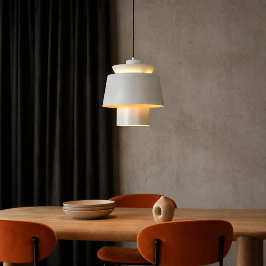 A modern multi-tiered pendant light with a white fabric shade and warm glowing bulbs, hanging above a wooden dining table with orange chairs in a contemporary room.