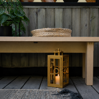 Modern gold metal outdoor lantern with glass panels displayed beneath a wooden bench creating warm ambient lighting.