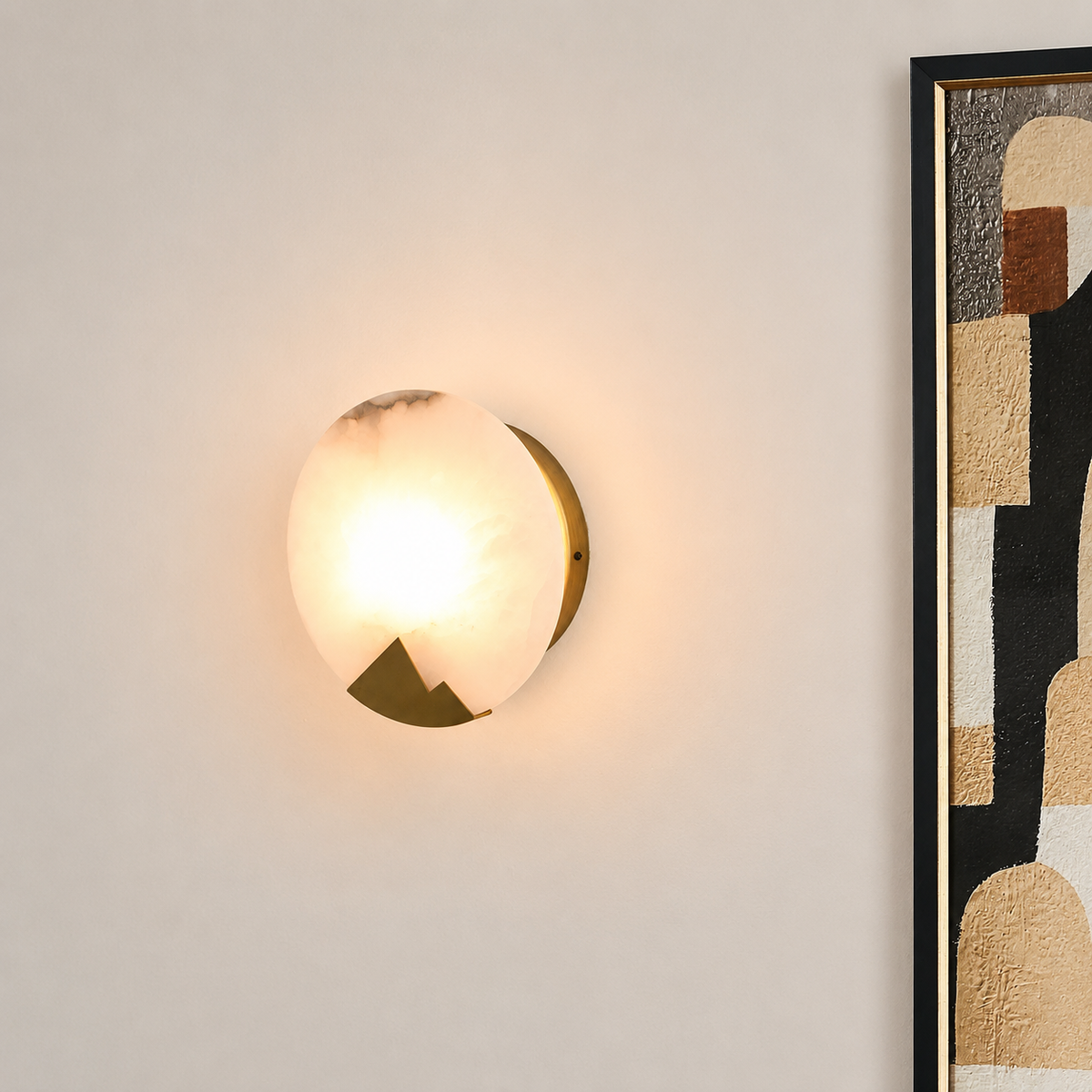 A modern wall sconce light fixture with a glowing frosted glass globe and a gold-toned geometric metal base, mounted on a light beige wall next to a framed abstract art print.