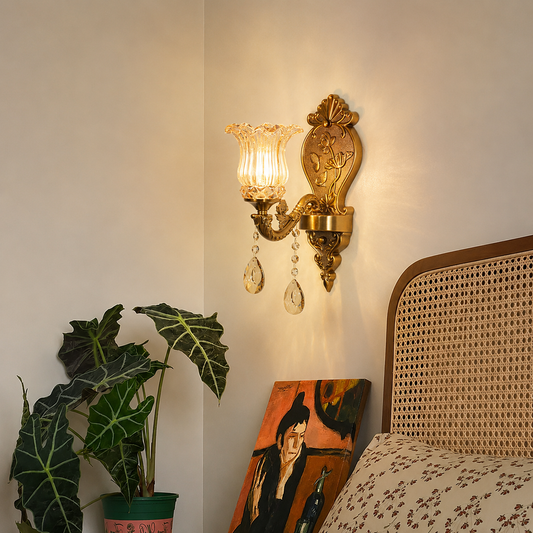 An ornate gold wall sconce with a fluted clear glass shade and hanging crystal drops, mounted on a beige wall in a styled bedroom corner next to a potted plant and a bed.