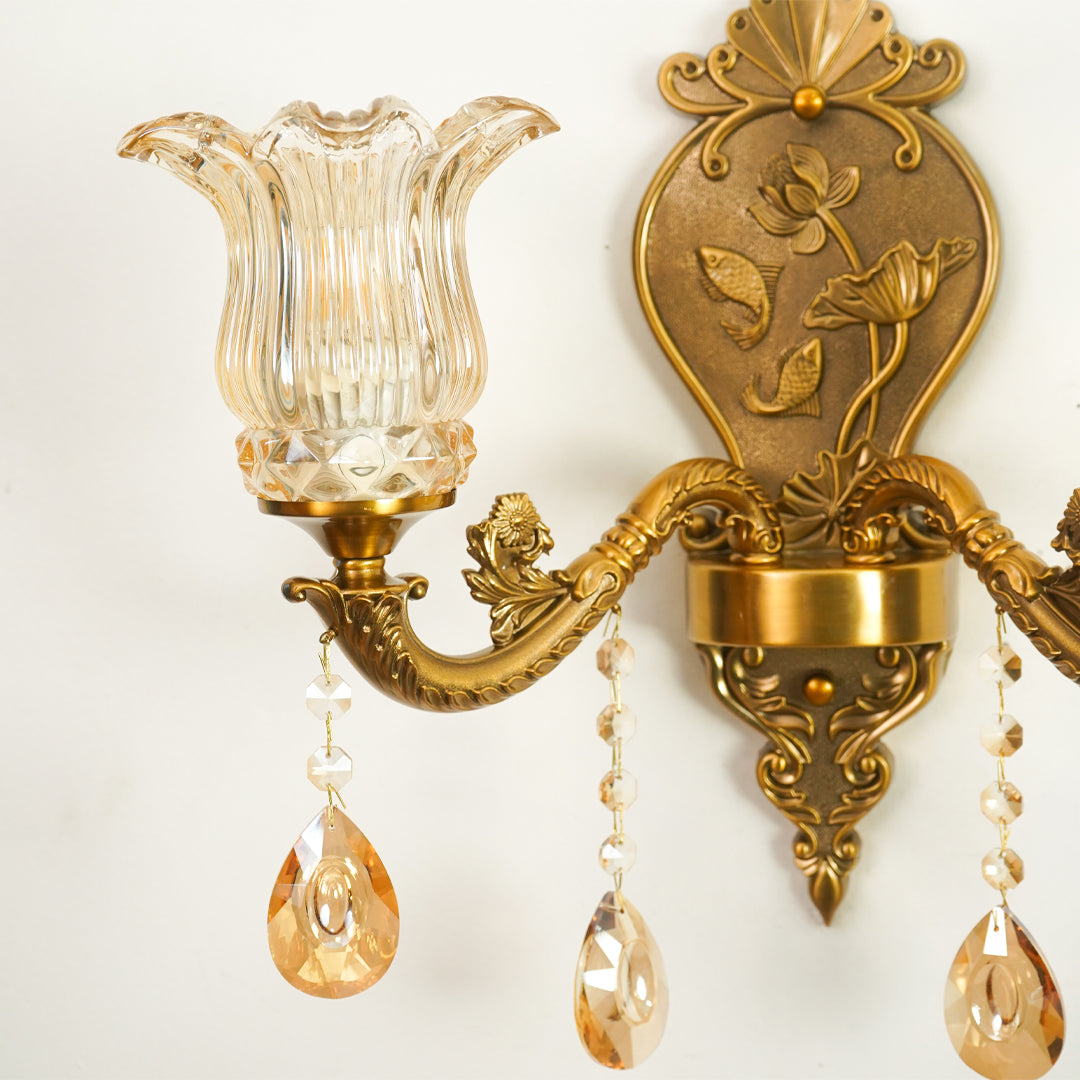 An ornate wall sconce with a fluted clear glass shade, antique gold/bronze metalwork, and hanging amber-tinted teardrop crystal drops against a plain light background.