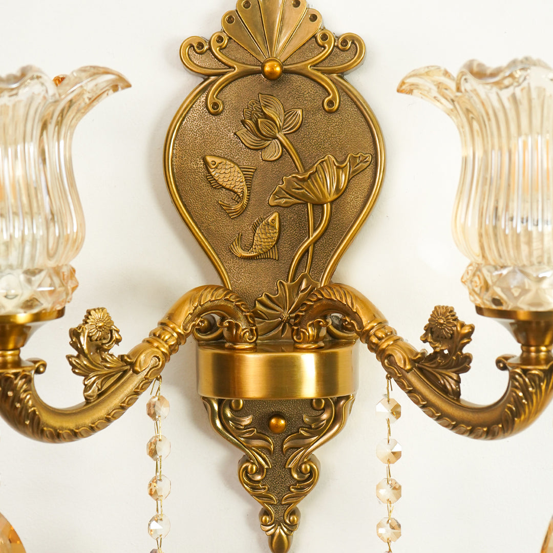 An ornate wall-mounted two-light candle sconce with antique gold/bronze metal arms and a central decorative shield motif featuring lotus flowers, flanked by clear ribbed glass shades and hanging crystal beads.