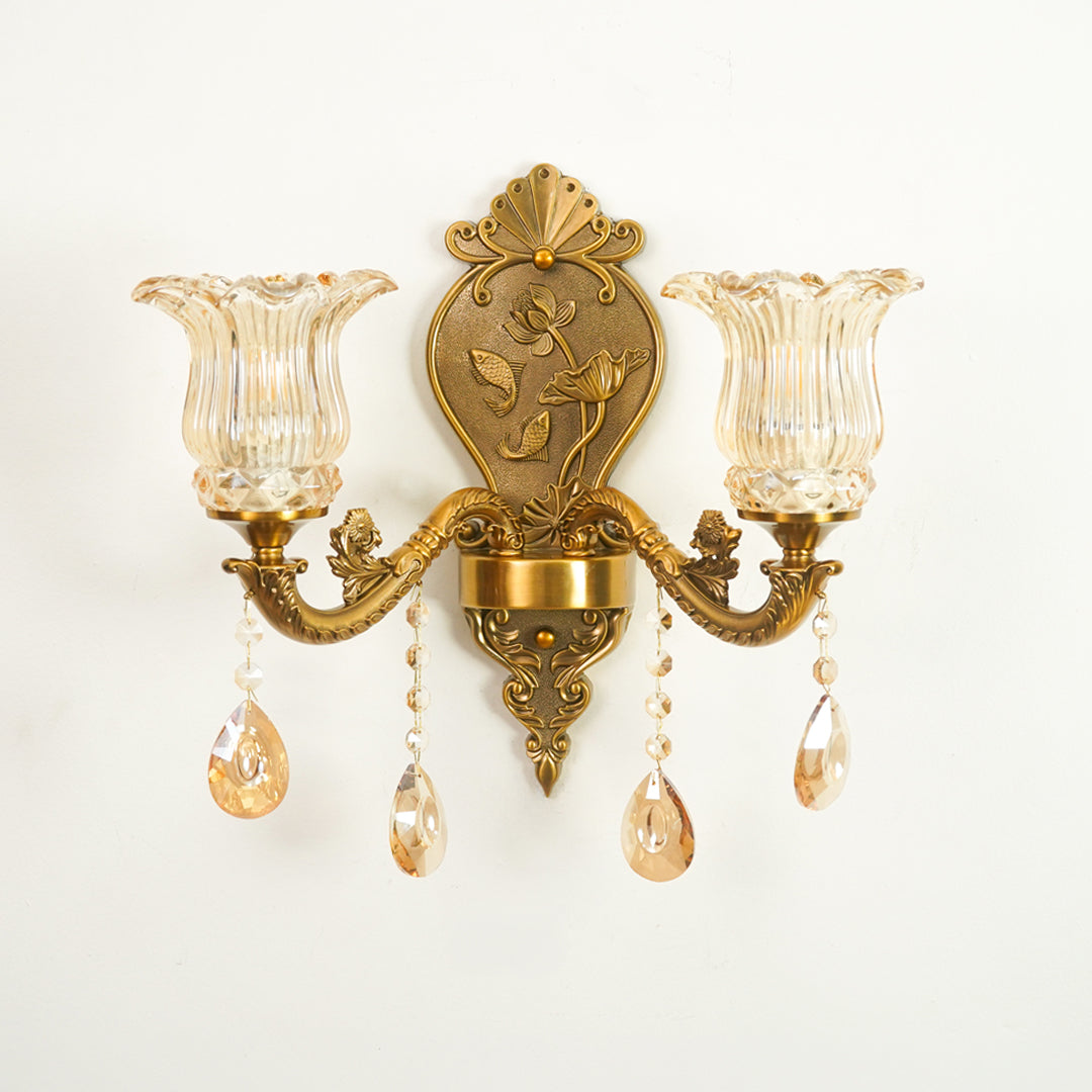 An ornate wall sconce with a gold-toned metal frame, two fluted clear glass shades, and hanging crystal teardrop pendants against a plain white background.