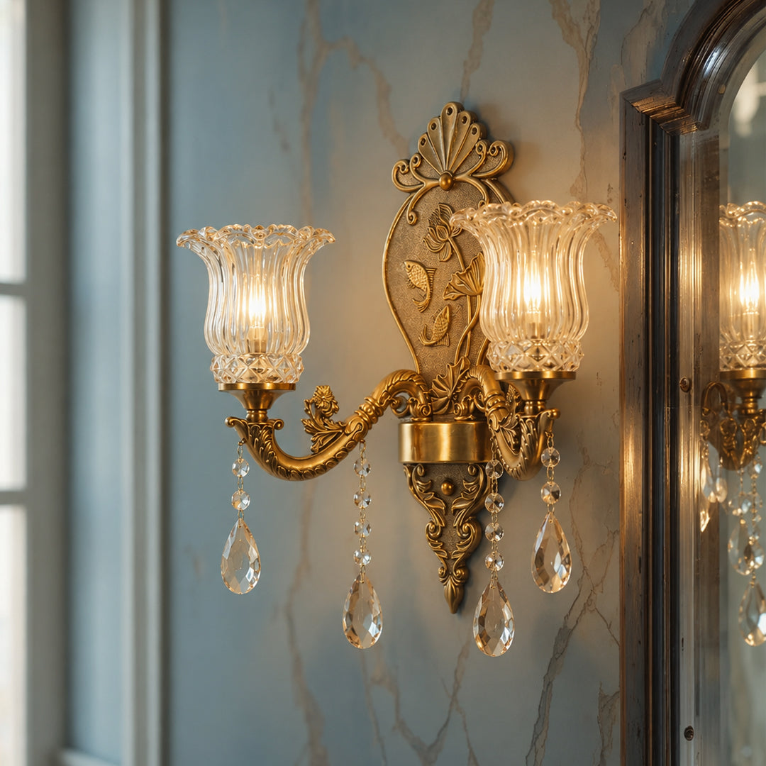 An ornate gold wall sconce with two fluted clear glass shades and hanging crystal teardrop pendants, mounted on a blue-gray marble-patterned wall next to a mirror.