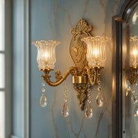 An ornate gold wall sconce with two fluted clear glass shades and hanging crystal teardrop pendants, mounted on a blue-gray marble-patterned wall next to a mirror.