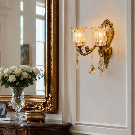 An ornate wall sconce with two ribbed glass shades, warm glowing bulbs, and hanging crystal drops, mounted on a white paneled wall next to a large gold-framed mirror.