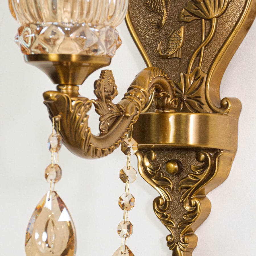 Closeup of antique gold wall light showing floral metal detailing and amber glass shade with crystal drops for refined interiors.