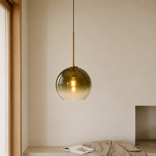 A modern pendant light with a translucent amber-green glass globe shade and a brass-colored rod, hanging in a minimalist beige room.
