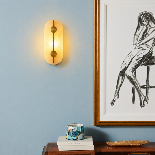 A modern wall sconce with a warm glowing oval glass shade and a vertical brass rod, mounted on a light blue wall next to a framed black-and-white sketch.