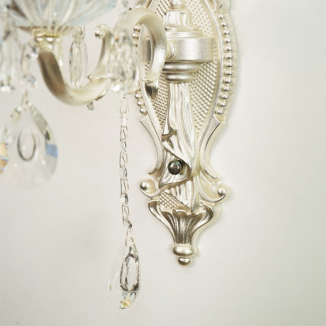 Close-up of an ornate wall sconce with a silver-toned metal frame, decorative scrollwork, and hanging clear crystal teardrop drops against a plain light background.