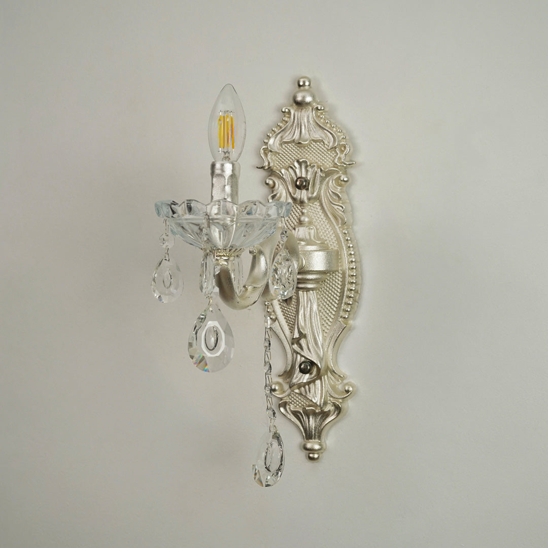 An ornate silver wall sconce with clear crystal teardrop pendants and a candle-style bulb, mounted on a plain light gray wall.