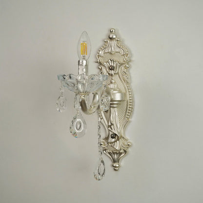 An ornate silver wall sconce with clear crystal teardrop pendants and a candle-style bulb, mounted on a plain light gray wall.