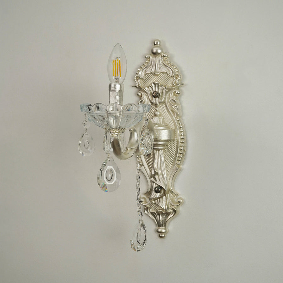 An ornate silver wall sconce with clear crystal teardrop pendants and a candle-style bulb, mounted on a plain light gray wall.
