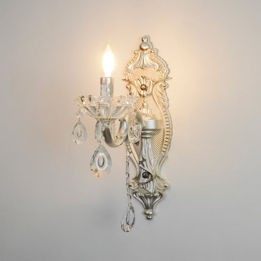 An ornate wall sconce with a silver-toned metal frame, clear crystal hanging drops, and a lit candle-style bulb, mounted against a plain light gray wall.